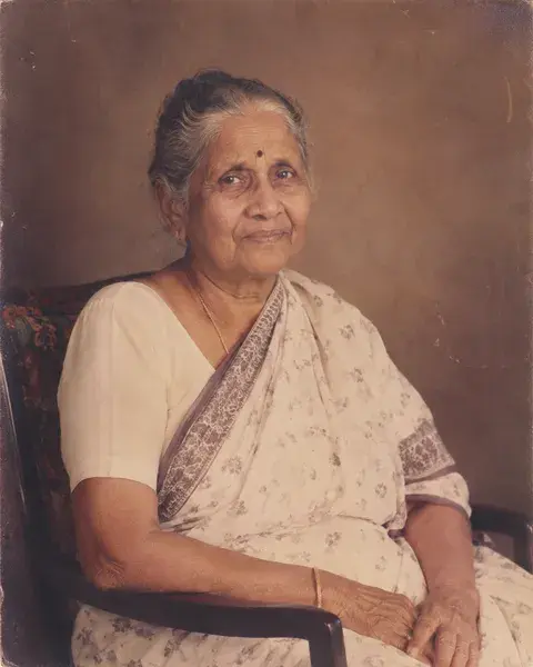 An Indian grandmother in her eighties with medium brown skin, grey hair in a neat bun and a simple cotton saree, photographed in the style of a carefully restored 1970s family studio portrait, retain age, natural grain and period clothing
