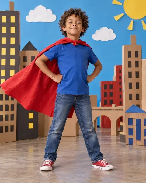 A seven-year-old boy with light brown skin and curly hair wearing a homemade red cape over a plain blue T-shirt and jeans, hands on hips in front of a playful cardboard city set, no branded superhero symbols