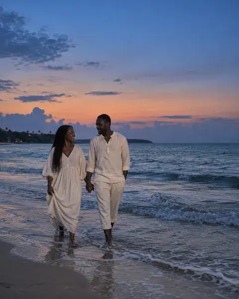 A dark-skinned African couple in their thirties, wearing loose off-white cotton clothing, walking barefoot along a quiet beach with small waves around their ankles