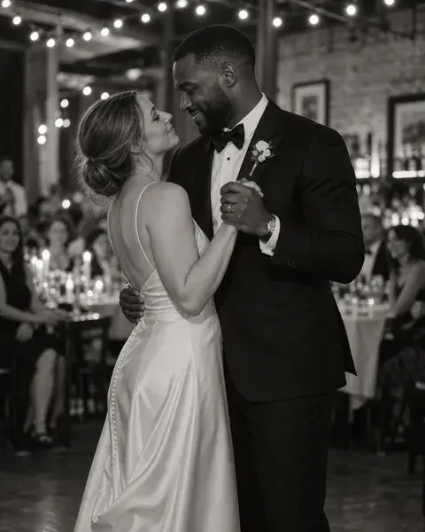 A white bride and Black groom in their thirties in a simple satin wedding dress and dark tuxedo, slowly dancing in an intimate restaurant wedding venue, guests softly out of focus