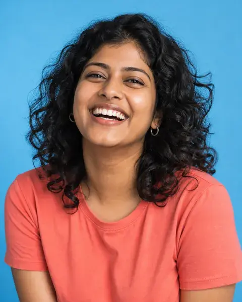 A young Indian woman with brown skin, curly shoulder-length hair and small silver hoops wearing a coral T-shirt against a saturated sky-blue backdrop, candid laughing expression