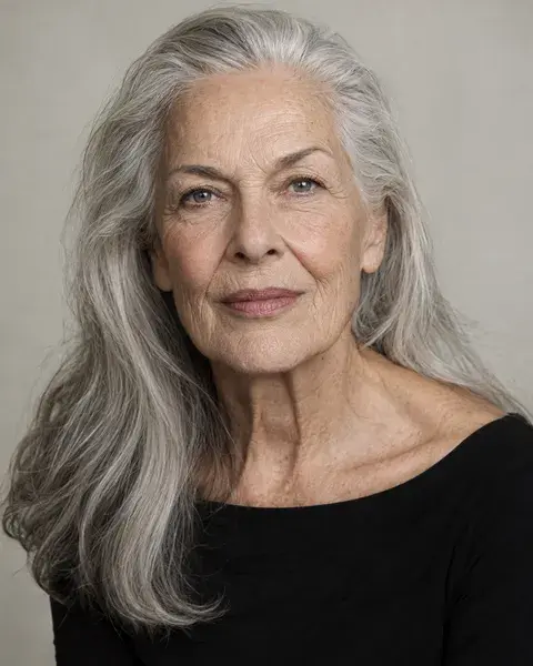 A woman in her seventies with fair skin, natural wrinkles and long silver hair wearing a black boat-neck top against a pale warm-grey studio backdrop, steady direct gaze
