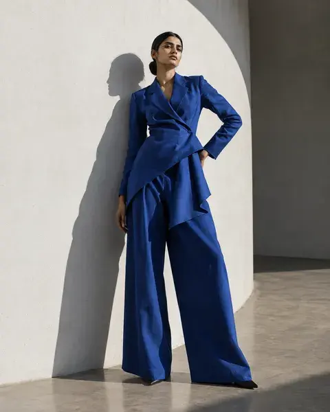 A tall South Asian woman with medium brown skin wearing a sculptural cobalt blue cotton suit, standing in a strong angular pose beside a curved white wall