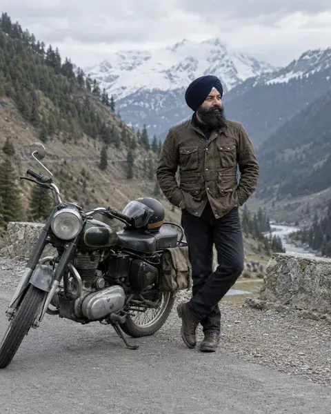 A Sikh man in his forties with a navy turban and full beard wearing a brown riding jacket, standing beside a parked classic motorcycle on a quiet mountain road, helmet resting on the seat