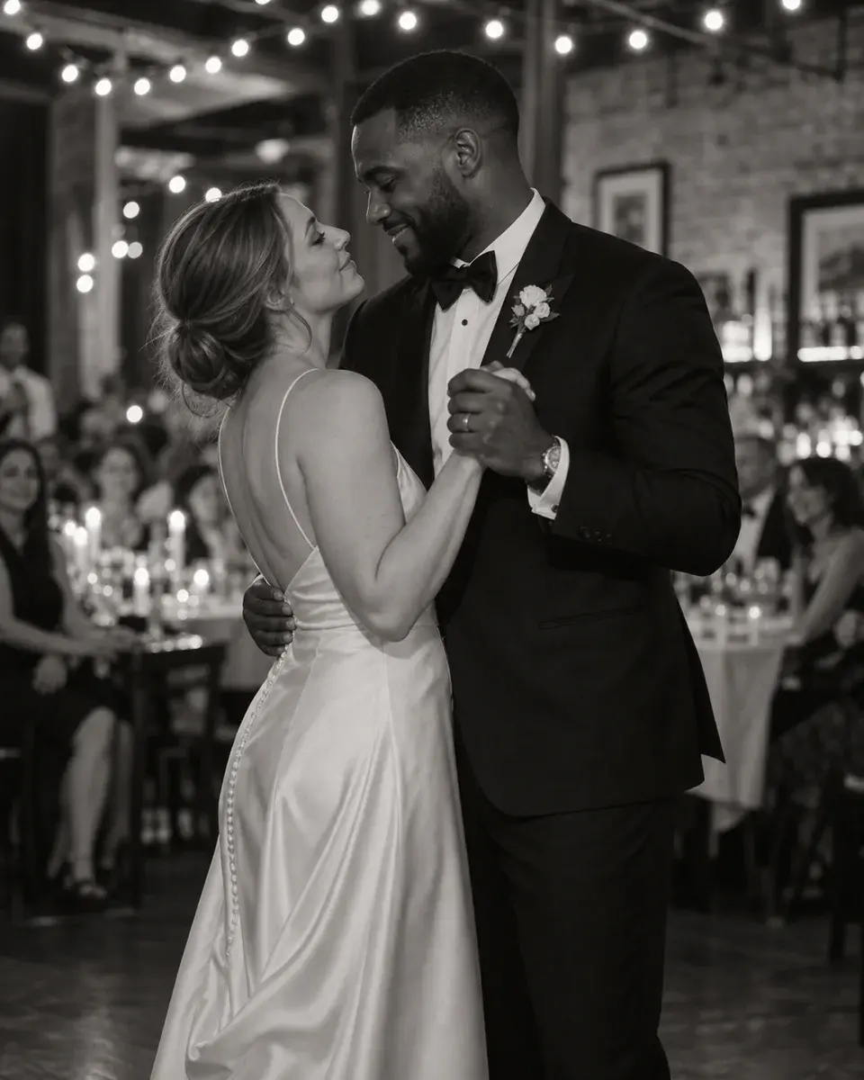 A white bride and Black groom in their thirties in a simple satin wedding dress and dark tuxedo, slowly dancing in an intimate restaurant wedding venue, guests softly out of focus