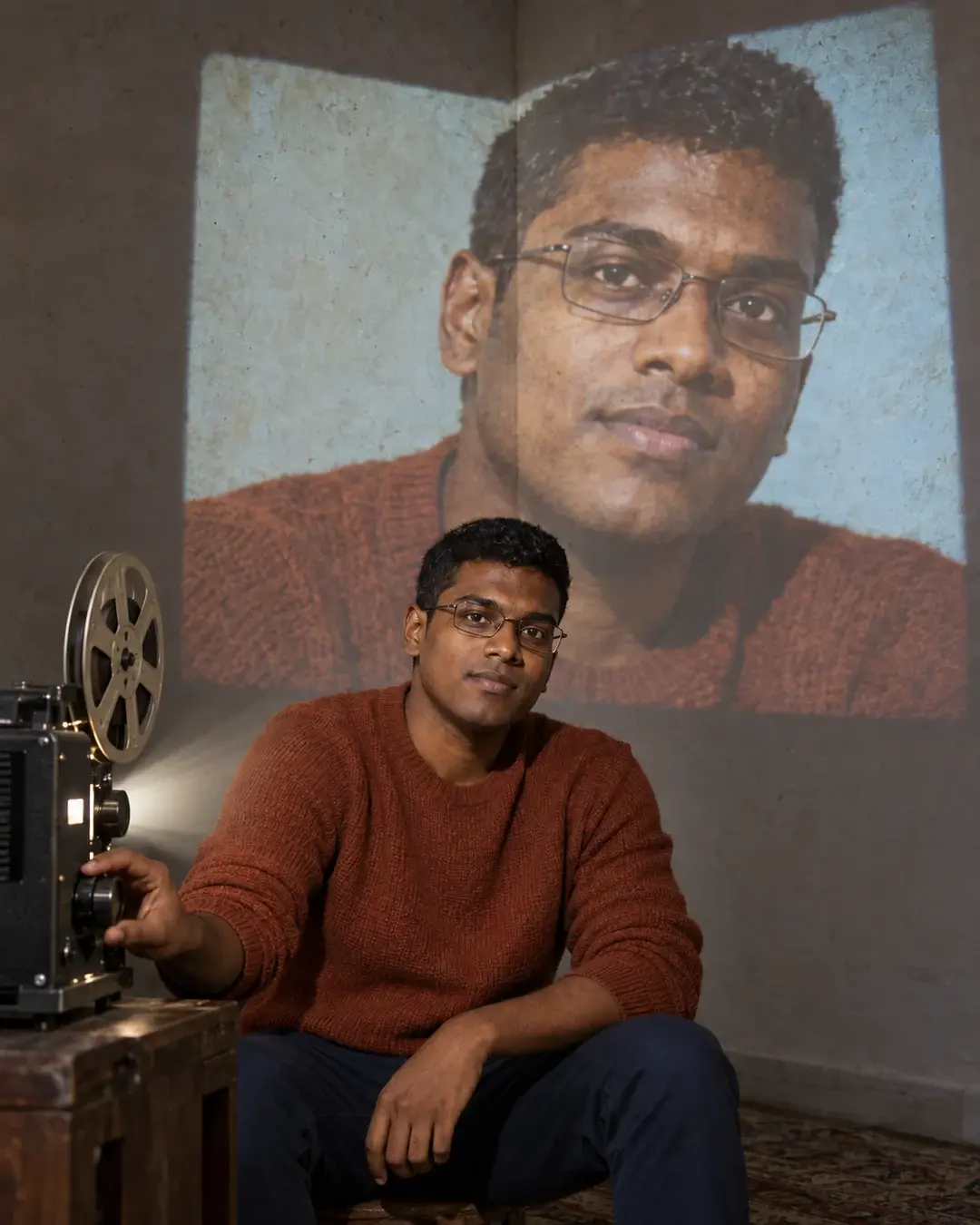 A seated man adjusts a projector beside a large projection of his own face across a plaster wall corner.